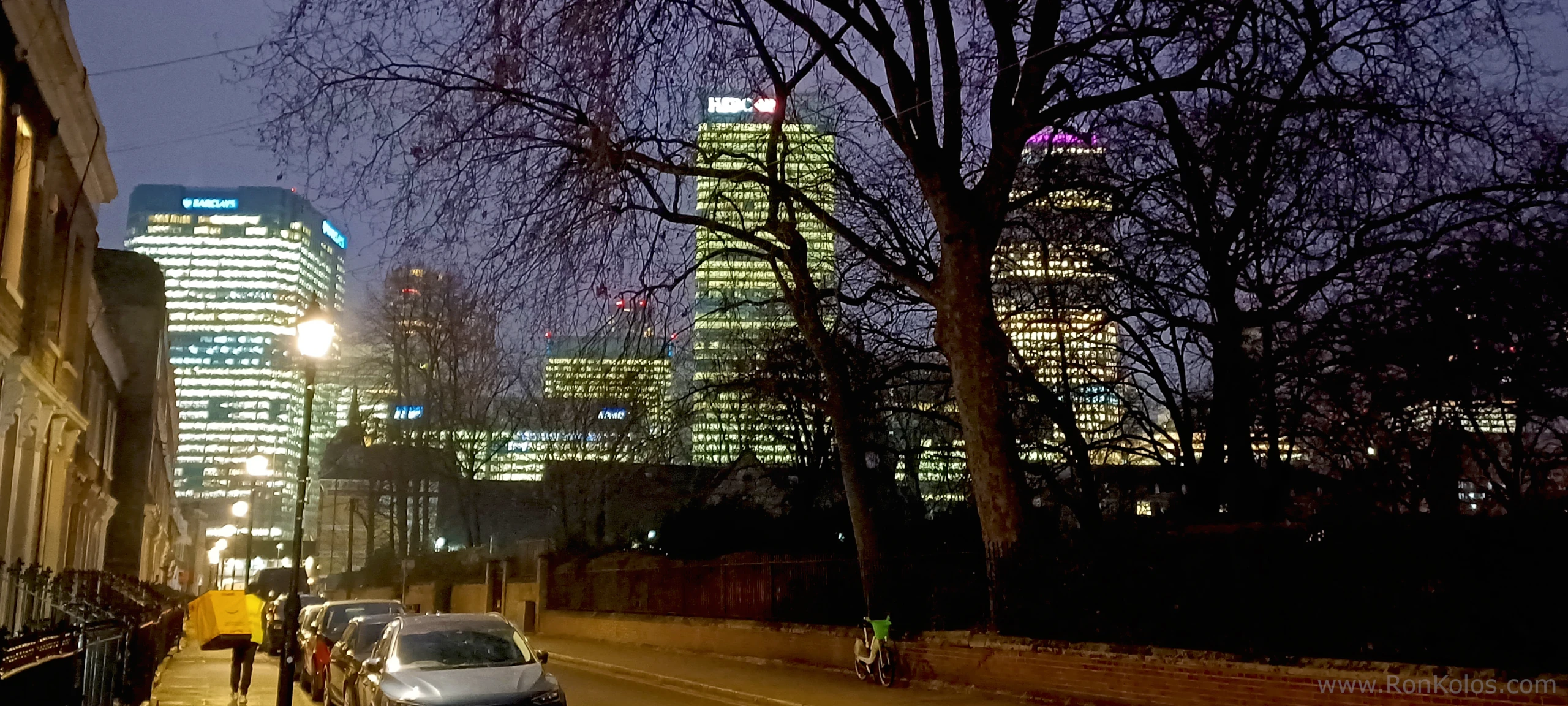 Photo of Uber or Deliveroo deliveryman walking down a dark street in London. Canary Wharf is in the background glowing bright.