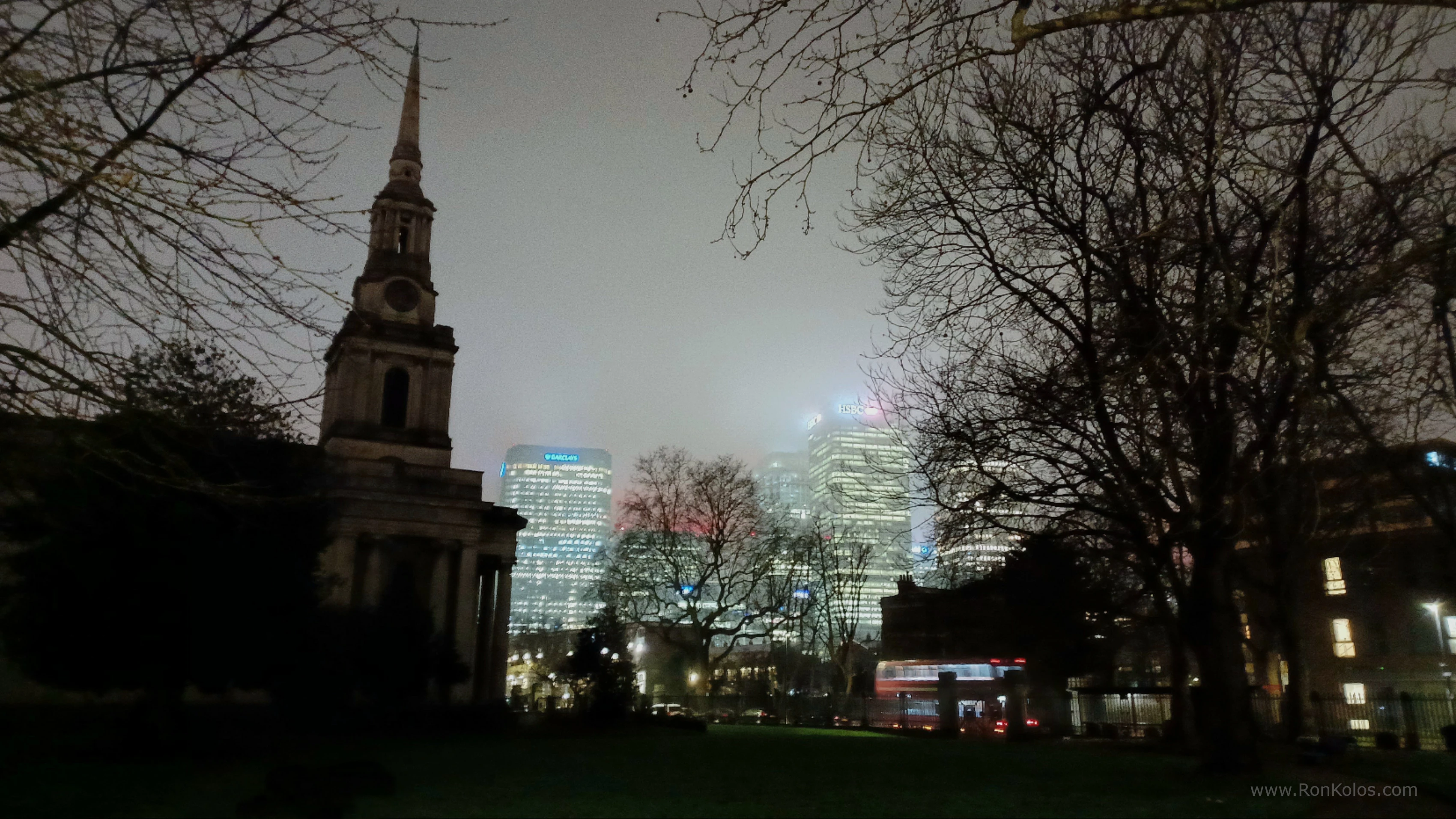 Photo of All Saints Church in Poplar, East London. Dark and eerie photo. Canary Wharf is glowing brightly through the mist and fog. Red London bus driving past.