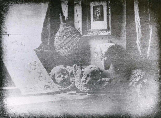 Image of the first every daguerreotype photograph. It shows some objects on a window sill. Some of them are dolls heads or maybe pieces of statues. There is also a picture handing on the wall and you can see a curtain.