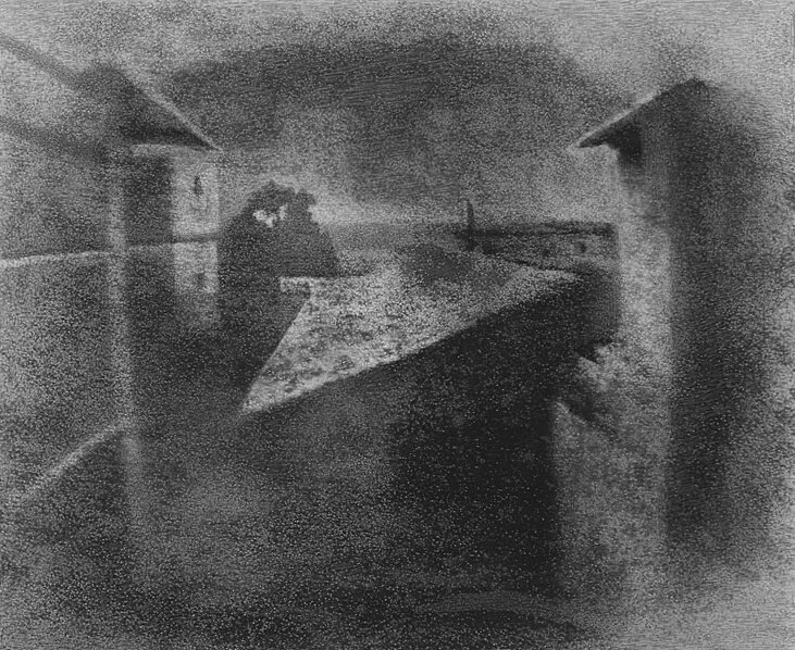 Image shows the first ever heliography photograph every taken. This is one of the first photos in human history. It shows a grainy image of a rooftop and it looks like something distorted and scratched onto a slab of metal or stone perhaps.