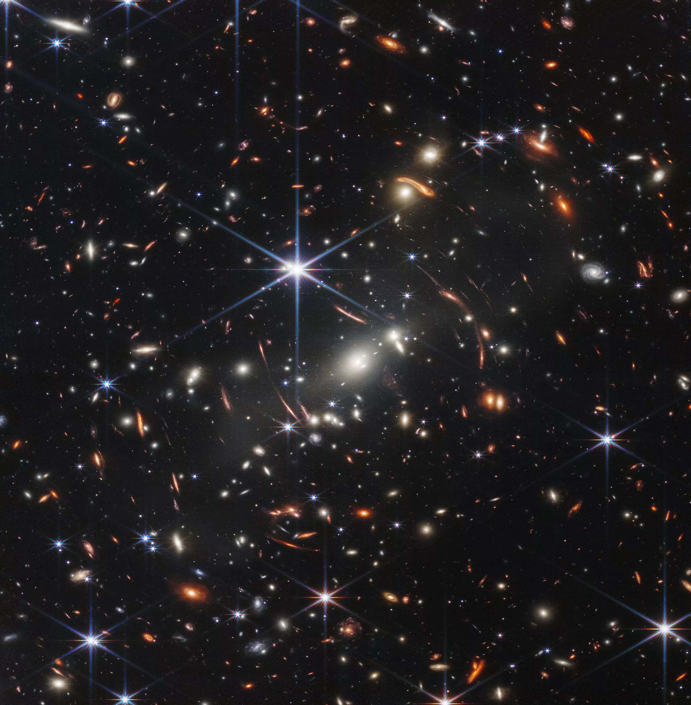 James Webb Space Telescope photograph of the first deep space image of distant galaxies going back to the very beginnings of the universe. A truly beautiful image showing thousands upon thousands of stars and galaxies shining brightly, and there is a huge galaxy cluster so big that the galaxies surrounding it are warped out of shape because of its gravitational pull. The warped galaxies look stretched and elongated, and the light from them is bent and magnified by the galaxy cluster, creating a lens effect.