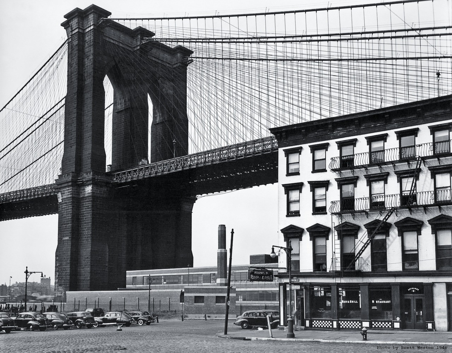 Photo of Brooklyn Bridge taken from the lower streets below. The photo is vintage. It is black and white.