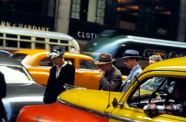Photo from the 1950s showing people crossing the street. There are many cars and its busy. There is an iconic New York taxi in the foreground. Taxis were colored red and yellow in those days.