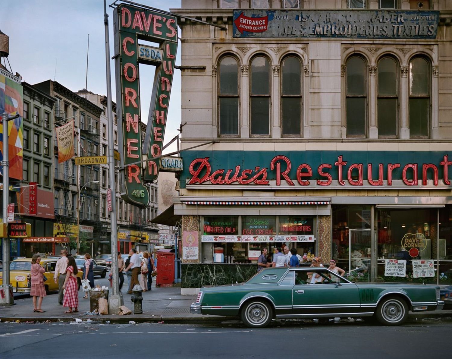 A groovy looking old photo of New York in the 70s. There is a cool car and groovy funky looking people, neon lights, busy streets.